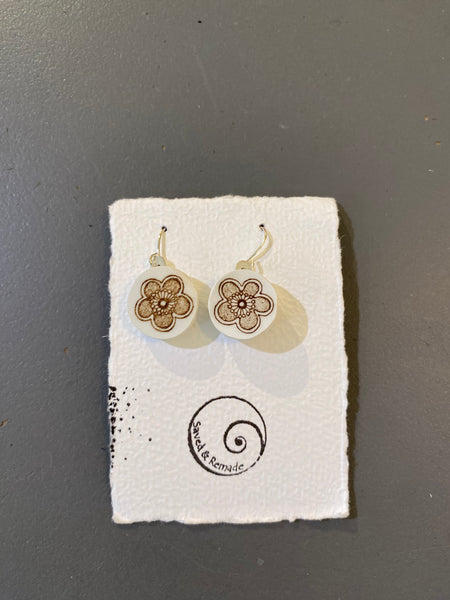 A pair of Saved & remade earrings by BUTIK KUBIK—white silverplated with brown floral designs, made from recycled porcelain, shown on a textured white card with spiral logo and Saved and Remade text, set against a gray background.