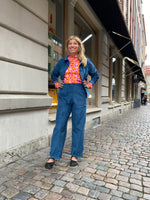 A woman stands confidently on a cobblestone street wearing Kubik corduroy trousers from BUTIK KUBIK, a blue jacket, black shoes, and a vibrant red and pink patterned top. She smiles with hands on her hips in front of large windowed buildings.