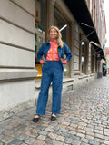 A woman stands confidently on a cobblestone street wearing Kubik corduroy trousers from BUTIK KUBIK, a blue jacket, black shoes, and a vibrant red and pink patterned top. She smiles with hands on her hips in front of large windowed buildings.