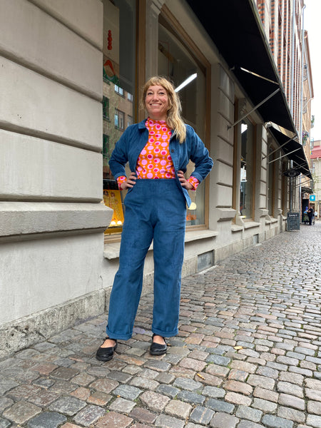 A woman stands confidently on a cobblestone street wearing Kubik corduroy trousers from BUTIK KUBIK, a blue jacket, black shoes, and a vibrant red and pink patterned top. She smiles with hands on her hips in front of large windowed buildings.