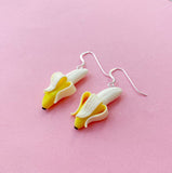 A pair of Becca design Banan earrings by BUTIK KUBIK, shaped like peeled bananas with sterling silver hooks, beautifully displayed on a pink background.
