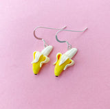 Two Becca design Banan earrings by BUTIK KUBIK, handgjorda och utformade som skalade bananer med örkrokar i sterling silver, visas mot en pastellrosa bakgrund.