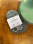 A pair of Essmoo earrings by BUTIK KUBIK, crafted from printed fabric, rests on a branded card atop smooth gray stones next to a green ceramic piece on wood. Nickel tested for your comfort.