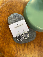 A pair of Essmoo earrings by BUTIK KUBIK, crafted from printed fabric, rests on a branded card atop smooth gray stones next to a green ceramic piece on wood. Nickel tested for your comfort.
