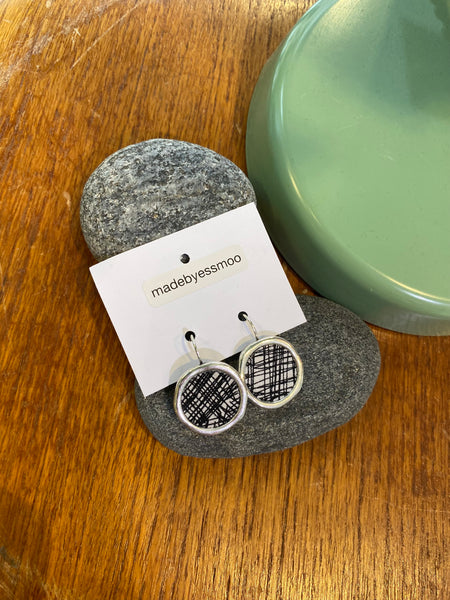 A pair of Essmoo earrings by BUTIK KUBIK, crafted from printed fabric, rests on a branded card atop smooth gray stones next to a green ceramic piece on wood. Nickel tested for your comfort.