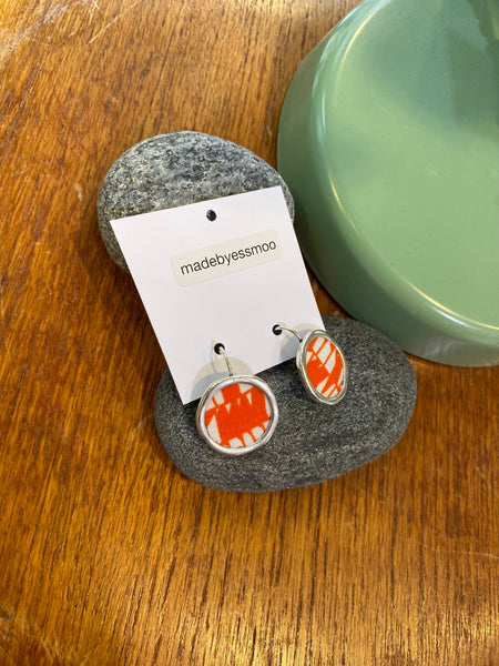A pair of silver-printed fabric Essmoo earrings with orange and white patterns by BUTIK KUBIK are displayed on smooth gray stones, with a madebyessmoo card behind them, all set on wood beside a green object.