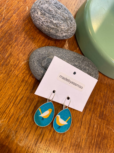 Essmoo earrings by BUTIK KUBIK: oval fabric earrings with blue backgrounds and yellow-and-white bird prints, displayed on a “madebyessmoo” card, rest on gray stones atop wood near a green object.