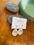 A pair of Essmoo earrings by BUTIK KUBIK—teardrop-shaped, gray cats flashing middle fingers—shown on a madebyessmoo card with smooth gray stones and a green lamp base. Nickel tested.