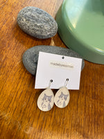 A pair of Essmoo earrings by BUTIK KUBIK—teardrop-shaped, gray cats flashing middle fingers—shown on a madebyessmoo card with smooth gray stones and a green lamp base. Nickel tested.