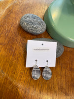 A pair of Essmoo earrings by BUTIK KUBIK, with a black and white grid fabric design, displayed on a branded card atop wood beside smooth grey stones and a green lamp base.