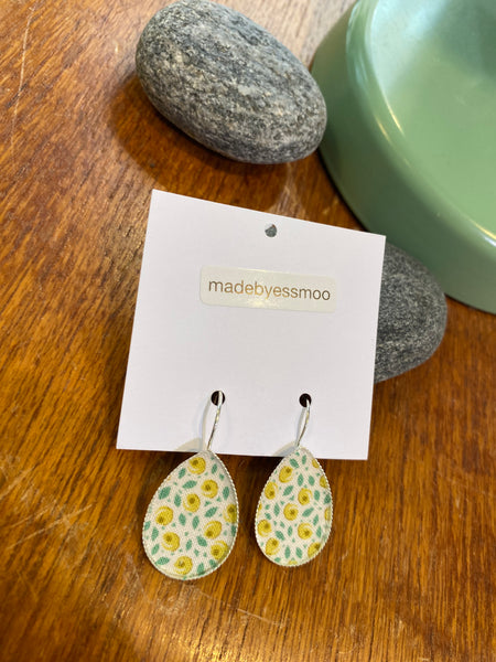 A pair of teardrop-shaped Essmoo earrings from BUTIK KUBIK, featuring a yellow and green floral pattern on printed fabric, are nickel tested and displayed on a white card resting on wood with grey stones and a green object nearby.