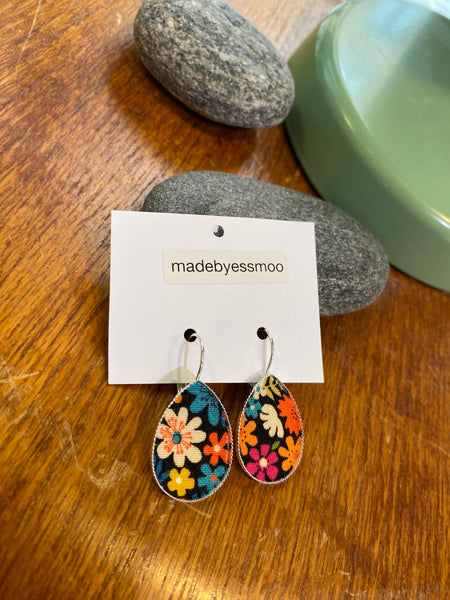 A pair of Essmoo earrings by BUTIK KUBIK featuring teardrop shapes and a colorful floral pattern are shown on a white madebyessmoo card, resting on wood with smooth gray stones and a green dish nearby.