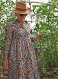 An older woman in a straw hat, sunglasses, and the Palava Tabatha floral dress by BUTIK KUBIK strolls through a lush greenhouse, gently touching tall tomato plants as sunlight filters through the leaves.