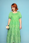A woman with short red hair wears the Cissi och Selma Ida klänning Plurr grön from BUTIK KUBIK, standing against a blue background, holding a small bouquet in her left hand while looking down and smiling slightly.