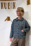 Wearing glasses and brown pants, a person stands by a wall with "KUBIK" and wooden hooks. They wear the BUTIK KUBIK Kubik Overshirt vintage patterned in deadstock fabric, smile slightly, and keep one hand in their pocket.