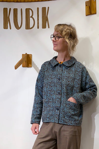 Wearing glasses and brown pants, a person stands by a wall with "KUBIK" and wooden hooks. They wear the BUTIK KUBIK Kubik Overshirt vintage patterned in deadstock fabric, smile slightly, and keep one hand in their pocket.