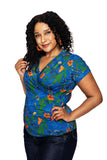 A woman with curly dark hair smiles, wearing the Cissi och Selma Jessica blouse Ogräs Blå from BUTIK KUBIK and matching orange earrings, standing with one hand on her hip against a white background.