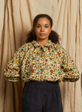 A woman with curly hair stands before beige fabric, wearing the BUTIK KUBIK Palava Joan Ivory Autumn Leaves Blouse—an autumnal top in green, red, and yellow—and dark high-waisted pants. She looks directly at the camera with a neutral expression.