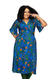 A woman with curly hair smiles, wearing the Cissi och Selma Josefine dress Ogräs Blå by BUTIK KUBIK. The blue floral omlottklänning in viskostrikå features fjärilsärmar. She poses with one hand on her hip and the other touching her hair against a white background.