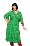 A smiling person with short black hair wears the Cissi och Selma Josefine dress Ängsblom Grön from BUTIK KUBIK, red tights, and has visible tattoos on one arm. They pose happily against a white background.