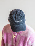 Someone in a BUTIK KUBIK Tropical Scandinavia cap TRUE WAY black, featuring faded stone-washed fabric, adjustable strap, and embroidered logo, pairs it with a pink and white checkered sweater while looking down to hide their face.