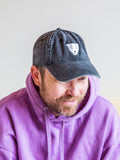 A bearded person in a BUTIK KUBIK Tropical Scandinavia cap black, featuring an ancient mask-inspired white logo, and a light purple hoodie, smiles and looks to the side against a plain light background.