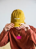 A person wearing the BUTIK KUBIK Tropical Scandinavia cap ochre tilts it downward, partially hiding their face. They pair it with an orange long-sleeve shirt and a necklace featuring pink COMMON NOISE COMMON VOICE letters.