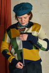 A person in a blue beret zips up the Palava Kate Mustard boxes cardigan from BUTIK KUBIK, featuring a colorful geometric pattern. A beige wall and red curtain serve as the background.