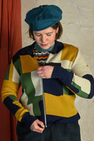 A person in a blue beret zips up the Palava Kate Mustard boxes cardigan from BUTIK KUBIK, featuring a colorful geometric pattern. A beige wall and red curtain serve as the background.