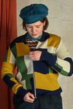 A person in a blue beret zips up the Palava Kate Mustard boxes cardigan from BUTIK KUBIK, featuring a colorful geometric pattern. A beige wall and red curtain serve as the background.