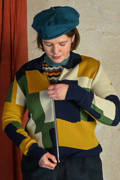 A person in a blue beret zips up the Palava Kate Mustard boxes cardigan from BUTIK KUBIK, featuring a colorful geometric pattern. A beige wall and red curtain serve as the background.