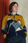A woman with short brown hair wears the BUTIK KUBIK Palava Kate Mustard peak zip-up cardigan, featuring a colorful mountain design. She stands with her hands in her pockets, gazing to the side before a beige wall and rust-colored curtain.