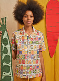 Wearing the BUTIK KUBIK Palava Kit pickle jars unisex summer shirt, made from a cotton-linen blend and featuring canned goods, a person with a natural afro poses by yellow backdrop holding a green prop and standing near a red carrot-shaped display.