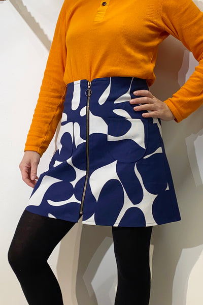A person poses in a bright orange long-sleeve top and the BUTIK KUBIK Kubik zip skirt nr 1 Vintage blue/white, featuring an abstract 70s pattern and front zipper, paired with black tights.