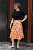 A barefoot person stands on stone slabs before dark wooden doors, wearing a black short-sleeve top and the BUTIK KUBIK Kubik skirt Twist Midi retro 1966 with orange, yellow, and pink florals, looking to the side with one hand on their hip.