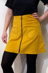 Wearing a black top and leggings, the person pairs them with BUTIK KUBIKs Kubik corduroy zip skirt nr 1 in senapsgult ökotex bomullsmanchester, featuring front pockets and a central zipper. Their hand rests confidently on their hip.