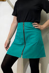 A person wearing a black short-sleeve top, black tights, and the BUTIK KUBIK Kubik zip skirt nr 1 Strömma Lotta grön in teal cotton with front zipper and two pockets stands with one hand on their hip against a light-colored wall.