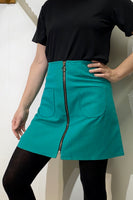 A person wearing a black short-sleeve top, black tights, and the BUTIK KUBIK Kubik zip skirt nr 1 Strömma Lotta grön in teal cotton with front zipper and two pockets stands with one hand on their hip against a light-colored wall.