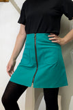 A person wearing a black short-sleeve top, black tights, and the BUTIK KUBIK Kubik zip skirt nr 1 Strömma Lotta grön in teal cotton with front zipper and two pockets stands with one hand on their hip against a light-colored wall.