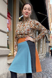 A woman stands on a cobblestone street wearing the BUTIK KUBIK Kubik skirt Twist short waffle multicolour (size M) and a floral blouse, smiling with one hand on her hip. City buildings are visible behind her.