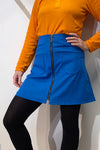 A person stands against a white wall wearing a bright orange top and the BUTIK KUBIK Kubik zip skirt nr 1 Strömma Lotta blå, featuring a front zipper and patch pockets. Their hand is on their hip, and black tights complete the look.