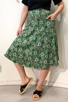 A person wears the BUTIK KUBIK Kubik skirt Twist Spring onion—a 50s-inspired green patterned skirt with pockets made from GOTS-certified fabric—paired with a black top and sandals, standing against a white wall with one hand in a pocket.