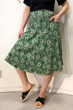 A person wears the BUTIK KUBIK Kubik skirt Twist Spring onion—a 50s-inspired green patterned skirt with pockets made from GOTS-certified fabric—paired with a black top and sandals, standing against a white wall with one hand in a pocket.