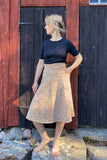A barefoot woman stands on rocks before a rustic wooden wall, wearing a black short-sleeve top and the BUTIK KUBIK Kubik skirt Twist Midi Radiokaka. She looks to the side, one hand on her hip.
