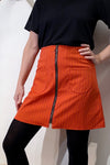 A person stands with one hand on their hip, dressed in a black t-shirt, black tights, and the BUTIK KUBIK Kubik zip skirt nr 1 Strömma Lotta röd—an orange-striped 70s-style skirt with a front zipper and two large pockets.