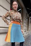 A woman smiles with one hand on her hip, wearing the BUTIK KUBIK Kubik skirt Twist short waffle multicolour size L and a patterned long-sleeve top, as she stands on a cobblestone street in front of a beige building.