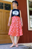 A barefoot person stands outside by a wooden red-and-yellow door, wearing a black cap, black graphic sleeveless shirt, and the BUTIK KUBIK Kubik skirt Twist Midi Molnblomma. They are looking to the side.
