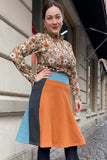 A woman smiles on a cobblestone street, wearing the BUTIK KUBIK Kubik skirt Twist short waffle multicolour size S with a floral blouse and black tights. Behind her is a stone building and shop windows.