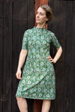 A woman poses confidently in front of a dark wooden wall, her light brown hair wind-swept, wearing the Kubik dress cinema midi vårlök by BUTIK KUBIK.