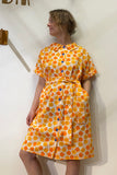 A woman stands against a white wall wearing the BUTIK KUBIK Kubik blouse dress vintage bluebell orange—an oversized, short-sleeved dress with a bold orange and white geometric pattern, blue buttons, and matching fabric belt.